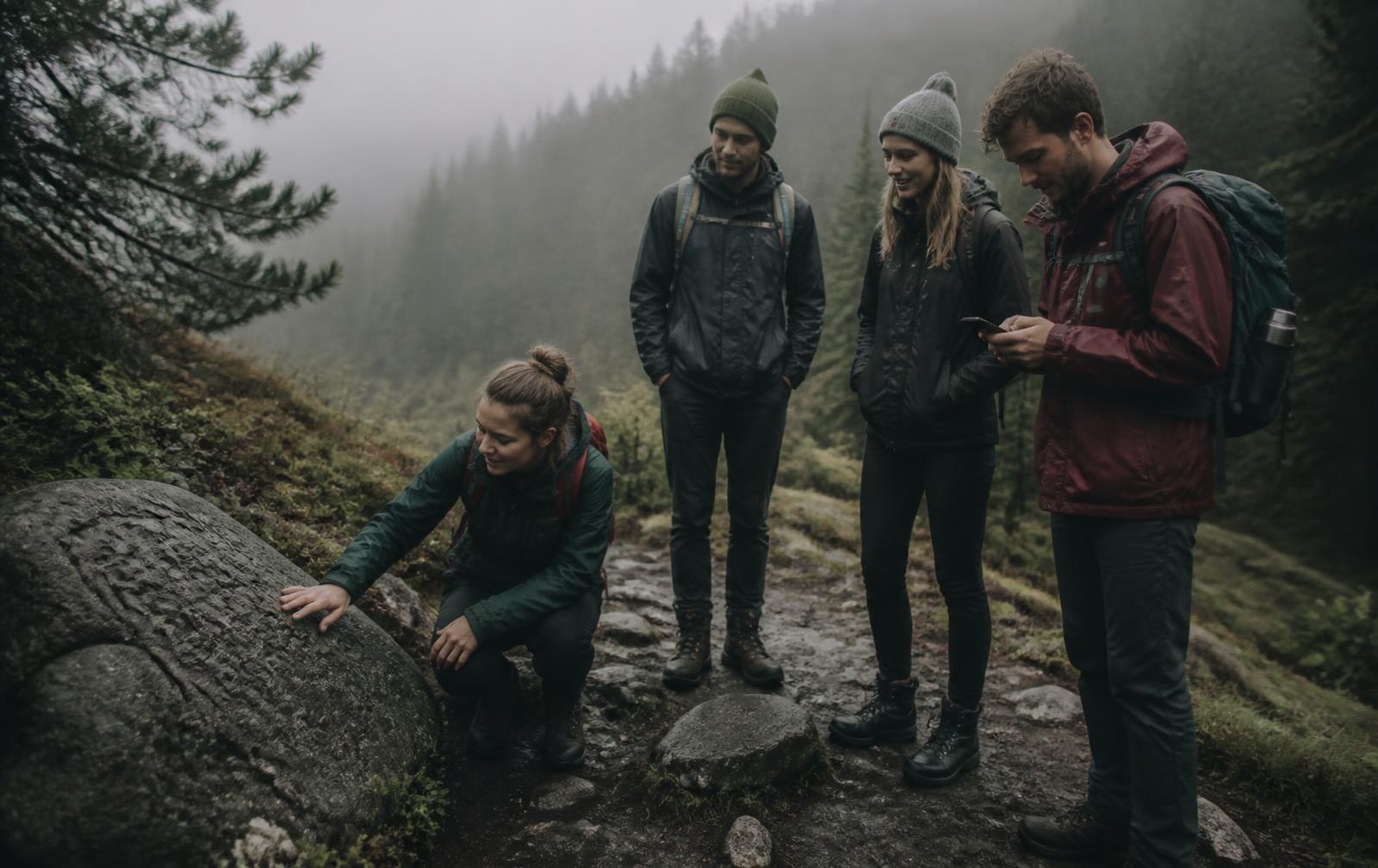 Skupina čtyř lidí v outdoorovém oblečení zkoumá kámen na mlžné horské cestě